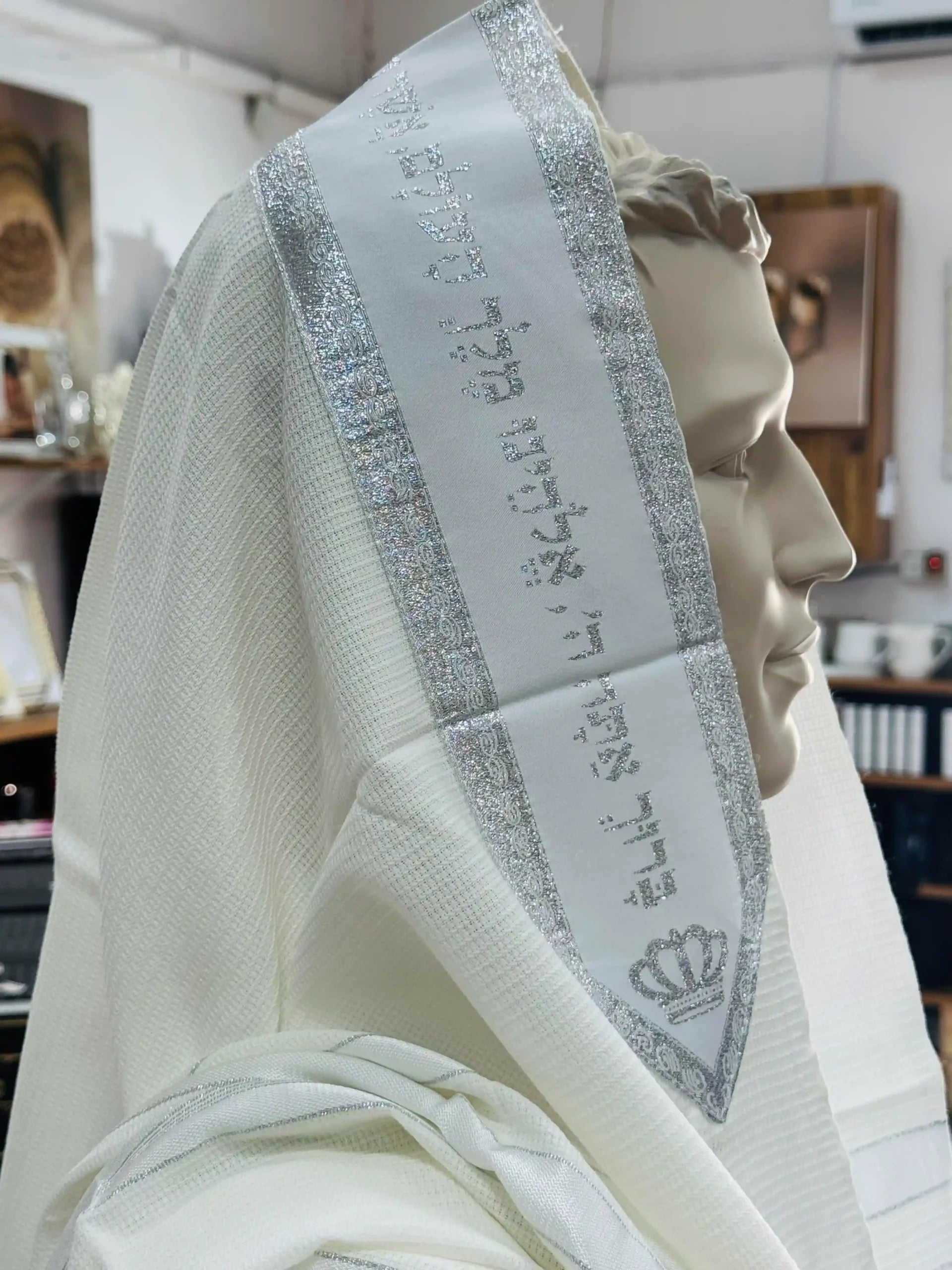 טלית לבנה מהודרת עם פס כסף ורקמת ברכת כהנים ועיטור כתר המונחת על ראש בובת תצוגה בתוך חנות יודאיקה