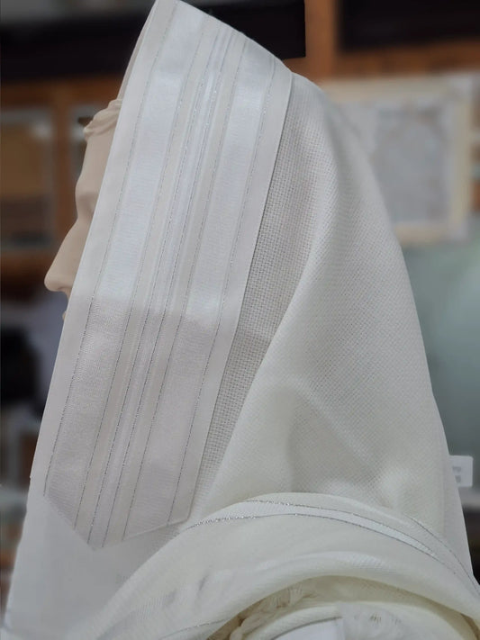 תקריב של טלית לבנה מהודרת עם פסי כסף דקים ורקמת קצה איכותית המונחת על ראש בובה לתצוגה