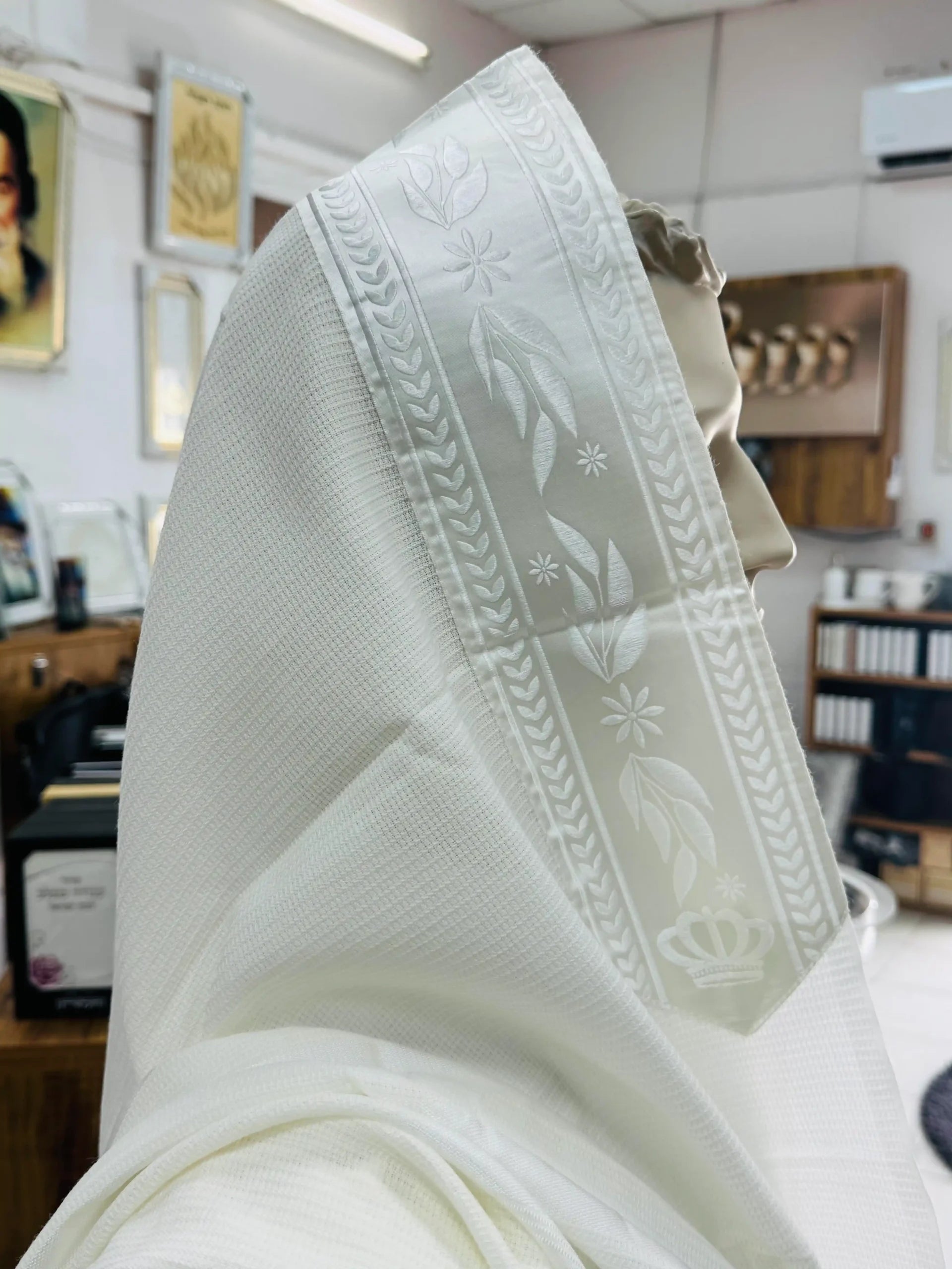 טלית לבנה עם פס דוגמת עלים ורקמה עדינה מונחת על ראש בובת תצוגה בתוך חנות יודאיקה Translation missing: he.content.product_image_main