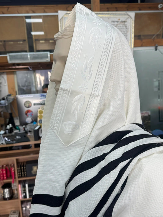 טלית לבנה עם פסי שחור ועיטור עלים ורקמת כתר עדינה המונחת על ראש בובת תצוגה בתוך חנות יודאיקה