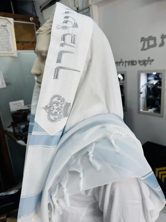 טלית לבנה לחתן בר מצווה עם פסים כחולים ורקמת כסף על קצה הטלית המונחת על ראש בובת תצוגה בתוך חנות יודאיקה