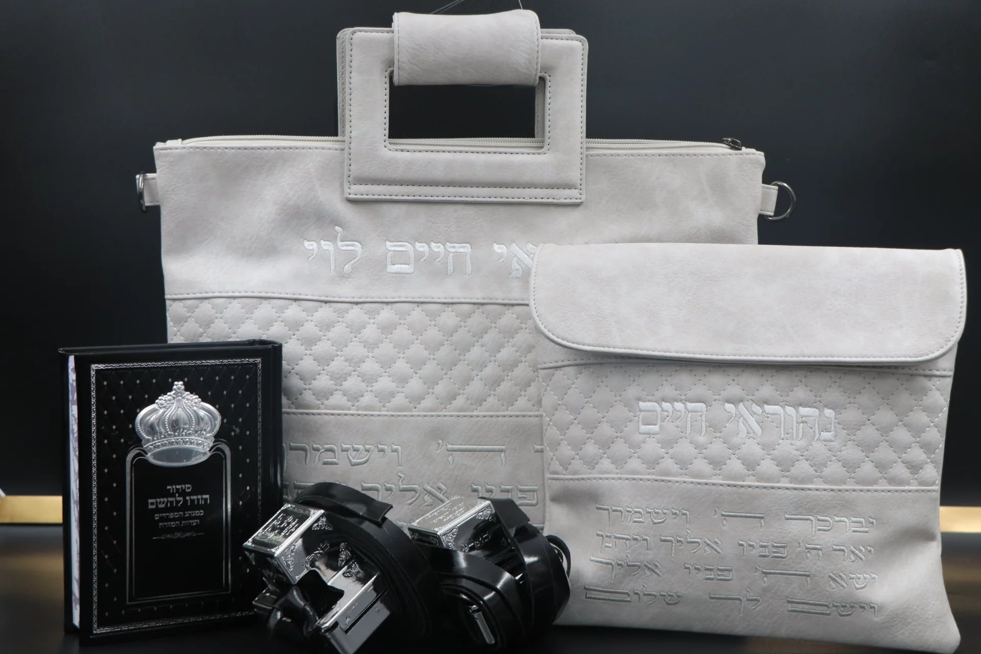 סט תפילין יוקרתי לבר מצווה הכולל תיקי טלית ותפילין לבנים בעיצוב קווילט עם רקמת שמות אישית בלבן, סידור שחור מהודר עם עיטור כתר ותפילין מהודרות על רקע כהה