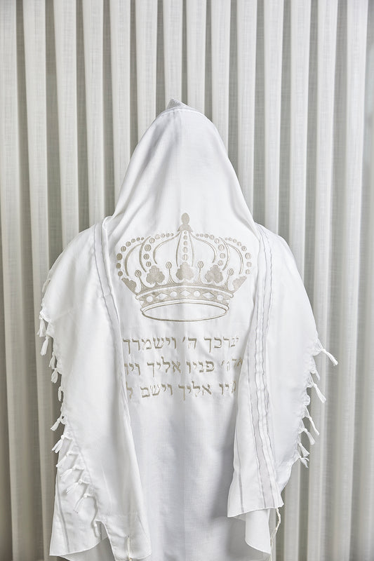 טלית לבנה עם רקמת כתר וכיתוב ברכת כהנים בגוון כסף, מוצגת על רקע וילון בהיר.