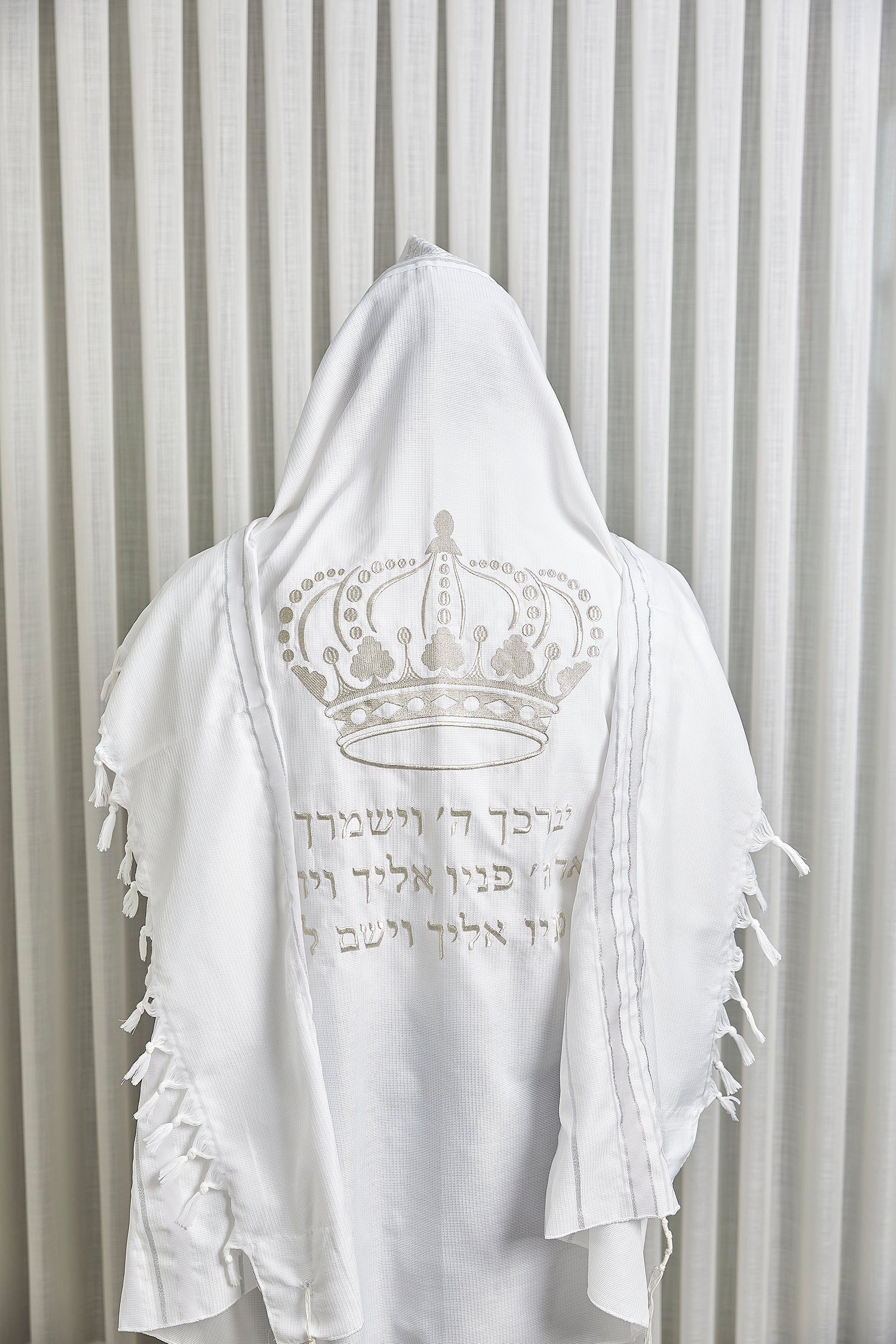 טלית לבנה עם רקמת כתר וכיתוב ברכת כהנים בגוון כסף, מוצגת על רקע וילון בהיר.
